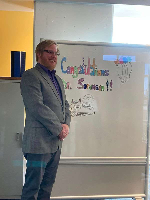 Lane Sorensen poses in front of a whiteboard that says "Congratulations Dr. Sorensen."