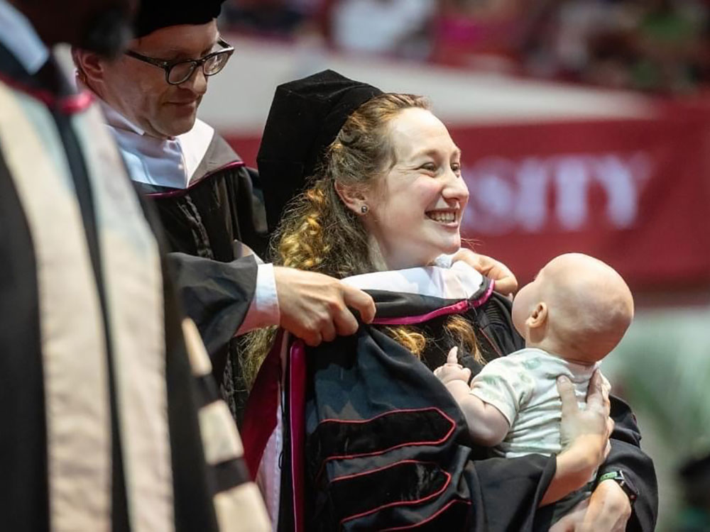 A graduate is hooded while holding a baby during a ceremony.